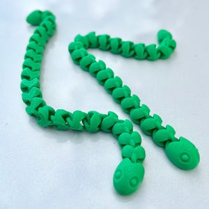 May include: Two bright green articulated snake toys with a segmented, flexible body design. Each snake features a rounded head with two circular indentations for eyes and a series of repeating, interlocking links that allow for fluid movement.