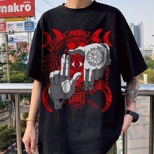May include: A black short-sleeve t-shirt featuring a large graphic print of two grey hands forming a frame shape. The design includes a red pentagram, a central eye, crescent moons, and the text Hellsing, Gate, and Aris in a circular pattern.