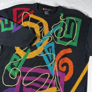 Può includere: Un maglione in maglia nero a maniche lunghe con un vivace motivo geometrico multicolore con forme rosse, gialle, verdi, viola e blu. Un sottile filo color oro è ricamato in una linea continua e sinuosa sulla parte anteriore del capo.