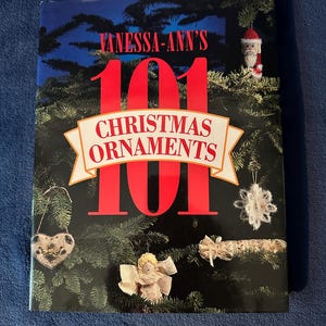 May include: A book titled Vanessa-Ann's 101 Christmas Ornaments with a cover featuring a dark green pine tree background decorated with various ornaments including a Santa figure, a white snowflake, a heart, an angel, and a fabric roll.