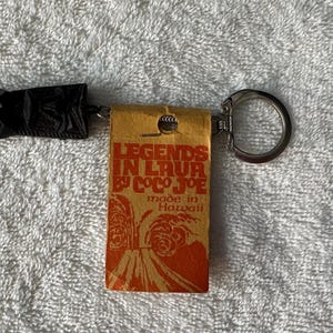 May include: A keychain featuring a black carved tiki figure attached to a metal ring. A rectangular orange tag is attached, printed with the text LEGENDS IN LAVA BY COCO JOE made in Hawaii, accompanied by a graphic illustration of a volcanic eruption.