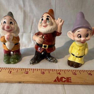 Può includere: Tre statuette di nani in ceramica posizionate dietro un righello di legno che misura fino a 17 pollici. La figura a sinistra indossa un cappello grigio, quella centrale un cappello marrone e tunica rossa, quella a destra un cappello viola e giallo.