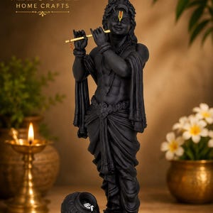 May include: A matte black statue of a figure playing a gold flute, featuring a yellow tilak on the forehead, draped clothing, and a small pot with white contents at the base. The base includes the text MurtiHome and the logo VELOURA HOME CRAFTS.