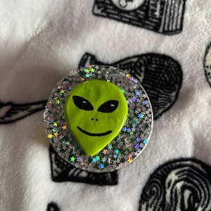May include: A circular phone grip featuring a bright green alien head with black eyes, nose, and mouth. The alien head is centered on a silver, glittery background filled with small, holographic star shapes that reflect light in various colors.