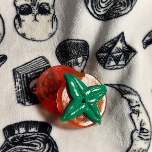 May include: A circular, translucent orange herb grinder featuring a prominent, three-dimensional green leaf-shaped topper. The grinder is positioned against a white fabric background patterned with black illustrations of skulls, crystals, and crescent moons.