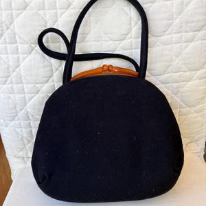 May include: A black handbag with a rounded silhouette and a single top handle. The bag features a distinctive amber-colored plastic clasp closure at the top, providing a contrast to the solid black fabric body of the accessory.