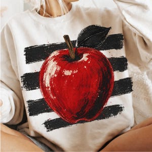 Puede incluir: Una sudadera de cuello redondo color crema con un gráfico grande y pictórico de una manzana roja con tallo marrón y hoja negra. La manzana está centrada sobre un fondo de cuatro franjas horizontales negras con efecto de pincelada.