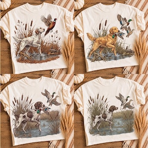 May include: A set of four off-white t-shirts, each featuring a detailed graphic illustration of a hunting dog in a marshland setting. The designs depict various breeds, including a spotted pointer and a golden retriever, interacting with ducks.