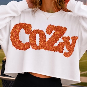 May include: A white long-sleeve cropped sweatshirt featuring the word Cozy printed in a textured orange and red autumn leaf pattern across the chest. The garment has a crew neckline and a raw, unfinished hemline at the bottom edge.
