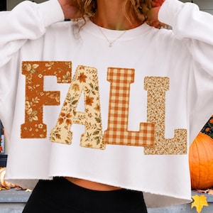 Op de afbeelding: Een wit sweatshirt met lange mouwen en een korte snit, voorzien van het woord FALL in grote blokletters op de borst. Elke letter heeft een eigen patroon: de F is bloemrijk, de A is bedrukt met bladeren, de L is oranje geblokt en de L is bloemrijk.