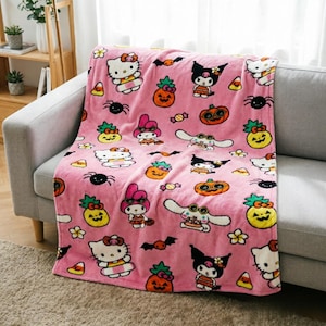 Puede incluir: Una manta de forro polar rosa suave con un patrón repetitivo de personajes de Sanrio, incluidos Hello Kitty, Kuromi, My Melody y Cinnamoroll, intercalados con piñas de calabaza naranjas, arañas negras, murciélagos y dulces de maíz.