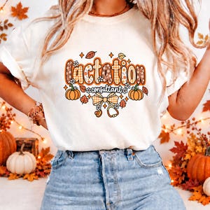 May include: A cream-colored short-sleeve t-shirt featuring a graphic with the text "lactation consultant" in orange and brown patterned letters. The design includes orange pumpkins, autumn leaves, a checkered bow, and a stethoscope icon.