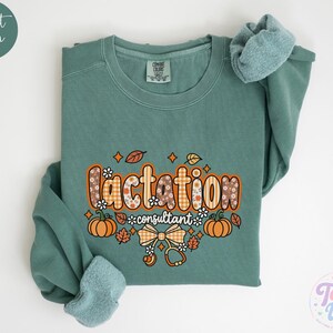May include: A light green crewneck sweatshirt featuring the text "lactation consultant" in a decorative font. The design includes orange pumpkins, autumn leaves, a plaid bow, and a stethoscope graphic with small white flowers.