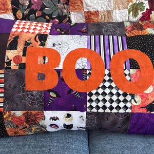 May include: A rectangular patchwork cushion featuring a variety of Halloween-themed fabric squares in orange, purple, black, and white. The word BOO is appliqued in large, textured orange letters across the centre of the quilted surface.