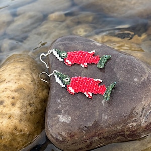 May include: A pair of dangle earrings crafted from small beads in the shape of fish. The fish have bright red bodies, white bellies, and dark green heads and tails. Each earring features a silver-toned metal hook for attachment to the ear.