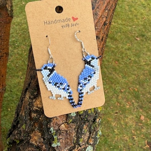 May include: A pair of beaded earrings shaped like blue jays, featuring shades of blue, white, and black glass beads. The earrings are attached to a rectangular tan cardboard display card with the printed text Handmade with love and a red heart.