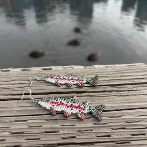 Op de afbeelding: Een paar visvormige oorbellen gemaakt van kleine, veelkleurige kralen. Het ontwerp is voorzien van een zilverkleurige haak, een witte buik, een roze zijstreep en een gespikkeld grijs en groen lichaamspatroon dat lijkt op een forel.