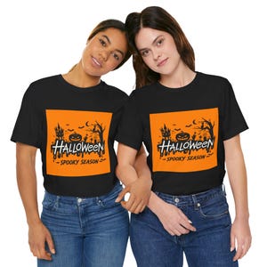 Peut inclure: Un t-shirt noir avec un motif rectangulaire orange sur la poitrine. Le motif présente une silhouette noire de maison hantée, une citrouille, un chat et des chauves-souris, avec le texte blanc "HALLOWEEN" et "SPOOKY SEASON" imprimé en dessous.