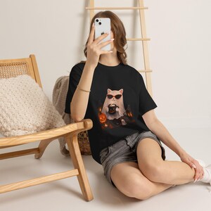 Peut inclure: Un t-shirt noir avec un motif représentant un chat blanc déguisé en fantôme, portant des lunettes de soleil noires et tenant une petite tasse noire avec un visage de citrouille. Le design inclut des citrouilles orange et des lanternes.