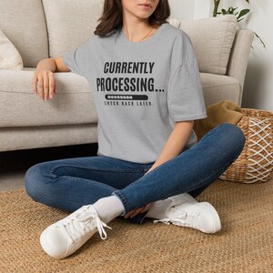 Op de afbeelding: Een grijs gemêleerd T-shirt met korte mouwen en de zwarte tekst "CURRENTLY PROCESSING..." boven een laadbalk en de woorden "CHECK BACK LATER." Het T-shirt wordt gedragen met een donkerblauwe spijkerbroek en witte veterschoenen.