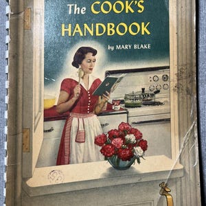 Könnte beinhalten: Ein spiralgebundenes Kochbuch mit dem Titel The COOK'S HANDBOOK von MARY BLAKE. Das Cover zeigt eine Illustration einer Person in einem roten Kleid und weißer Schürze, die einen Löffel und ein Buch in einer Küche mit Herd und Blumen hält.