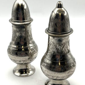 May include: Two silver-toned metal shakers with a bulbous body and a tapered neck. Each shaker features a removable dome-shaped lid with a pointed top and intricate etched geometric patterns covering the entire surface of the metal body.