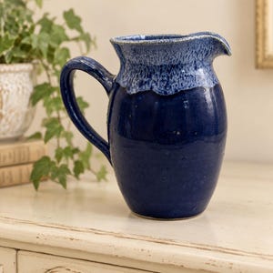 May include: A ceramic pitcher with a glossy deep blue finish on the body and a textured, lighter blue drip glaze around the rim. The vessel features a curved handle on the side and a prominent pouring spout, showcasing a handcrafted appearance.