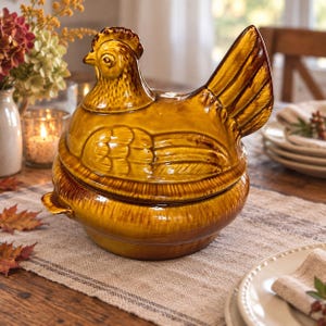 May include: A glossy, mustard-yellow ceramic chicken-shaped lidded casserole dish. The lid features a sculpted hen with detailed feathers and a raised tail, while the base is a round, deep bowl with small side handles, finished in a warm glaze.