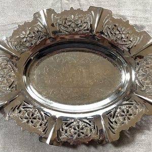 May include: An oval silver-toned metal tray featuring a polished centre with an etched floral pattern and a wide, scalloped rim decorated with intricate cut-out designs and embossed flower motifs around the entire perimeter of the dish.