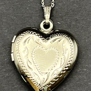 May include: A silver-toned heart-shaped locket pendant suspended from a matching silver-toned chain. The locket features an engraved decorative border with a central heart motif and a smooth, polished finish on the front surface.