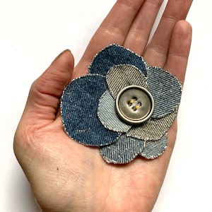 May include: A denim flower brooch with a silver button in the center. The flower is made of multiple layers of denim fabric with a glittery edge.