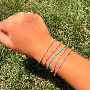 May include: A set of four thin beaded bracelets worn on a wrist. The bracelets feature small, round beads in alternating colors of bright pink, light blue, and metallic gold, arranged in a simple, delicate, and uniform circular pattern.