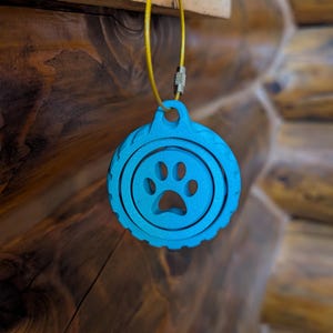 May include: A bright turquoise circular tag featuring a cutout paw print design in the centre. The tag has a textured, ridged outer edge and is suspended from a thin, flexible yellow metal cable loop with a silver-toned threaded barrel connector.