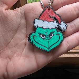 Op de afbeelding: Een 3D-geprinte sleutelhanger met een groen personage met een ondeugende uitdrukking, die een rood-witte kerstmuts draagt. Het ontwerp is zwart omlijnd en de sleutelhanger is bevestigd aan een zilverkleurige metalen ring en ketting.