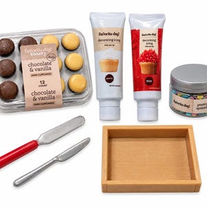 May include: A cupcake decorating kit including a clear plastic container of 12 chocolate and vanilla mini cupcakes, two tubes of Favorite Day decorating icing in white and red, a jar of pastel party sprinkle mix, two metal knives, and a wooden tray.