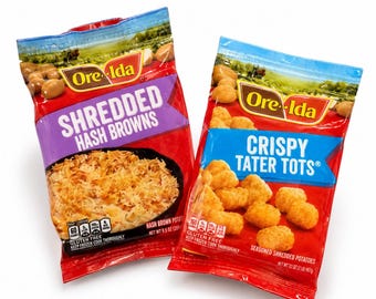 Op de afbeelding: Twee zakken met diepvries aardappelproducten van Ore-Ida. De linkerzak bevat Shredded Hash Browns met een paars label, en de rechterzak Crispy Tater Tots met een blauw label. Beide zakken tonen een landschapsafbeelding, logo en tekst.