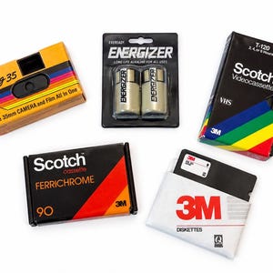 Puede incluir: Una colección de artículos multimedia y de energía vintage que incluye una cámara Kodak Fling 35 amarilla, un blíster de dos pilas Energizer, un videocasete VHS Scotch T-120 negro, un casete Scotch Ferrichrome 90 negro y un disquete 3M en funda blanca.
