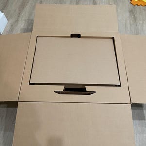 May include: A large, open, brown cardboard shipping box containing a rectangular cardboard insert. The insert features a central rectangular cutout and a smaller base cutout, designed to securely hold a flat-screen monitor or television.