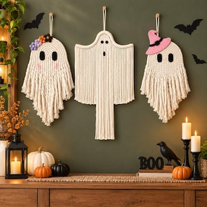 May include: Three macrame ghost wall hangings made of off-white cord. The left ghost features a crown of colorful pom-poms and pink cheeks. The center ghost has a simple face. The right ghost wears a pink felt witch hat. All have black felt eyes.