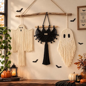 May include: Three macrame wall hangings suspended from a wooden branch. Two off-white ghost figures with black felt eyes and mouths flank a central black bat figure featuring two round wooden bead eyes and a long, thick central tassel.