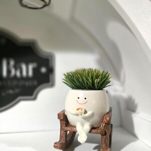 May include: A small, off-white, egg-shaped planter with a smiling face, holding a tiny cup, and sitting on a miniature brown wooden rocking chair. The planter contains a dense cluster of spiky, vibrant green artificial grass on top.