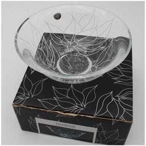 May include: A clear glass bowl featuring a delicate etched floral pattern and a circular logo on the base. The bowl rests on a black square box decorated with a matching white line-art floral design and a label reading Filigran Design Lin Utzon.