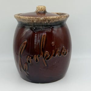 May include: A dark brown ceramic cookie jar with a glossy finish and a matching lid. The jar features the word Cookies embossed in a cursive script on the front. The rim of the jar and the lid edge have a textured, lighter brown drip glaze.