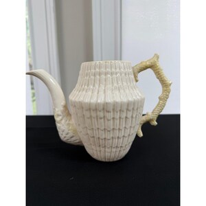 May include: A cream-colored ceramic teapot featuring a textured, ribbed body that mimics the appearance of a shell. The handle and spout are designed with a detailed, branch-like or coral-inspired texture, providing a distinct contrast to the main body.