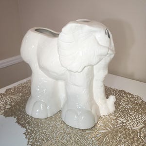 May include: A white ceramic elephant-shaped planter or vase with a glossy finish. The elephant is depicted in a standing position with its trunk slightly curled upward. The surface features subtle molded textures and black painted eye details.