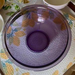 May include: A round, translucent purple glass bowl with a smooth, wide rim and a textured, pebbled base. The bowl features a deep, circular center indentation and a clear, polished finish that allows light to pass through its vibrant violet surface.