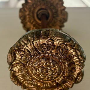 May include: A metallic gold-toned curtain tieback featuring a circular, ornate floral design with intricate embossed petal details. The hardware has a weathered, antiqued finish with a central cluster of small raised spheres and a long rod.