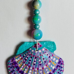 Könnte beinhalten: Ein hängendes Ornament mit einer bemalten Muschel in Violett-, Türkis- und Weißtönen mit einem Punktmuster. Die Muschel hängt an einer weißen Schnur, die mit drei runden, mehrfarbigen Perlen in Blau-, Grün- und Gelbtönen verziert ist.
