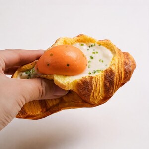 Può includere: Un croissant dorato farcito con una salsa bianca cremosa e un centro liscio di colore arancione brillante che ricorda un tuorlo d'uovo, guarnito con piccole scaglie di erbe verdi. La pasta ha una consistenza sfogliata e una forma curva.
