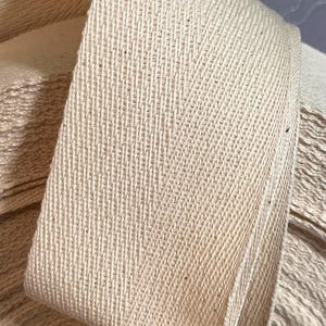 May include: Close-up of a beige cotton twill tape with a herringbone weave. The tape is folded and stacked, showing the texture of the fabric.